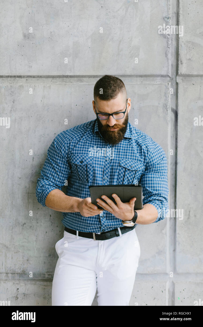 Tanga barbuto imprenditore indossando occhiali e plaid shirt appoggiata contro la parete utilizzando tablet Foto Stock