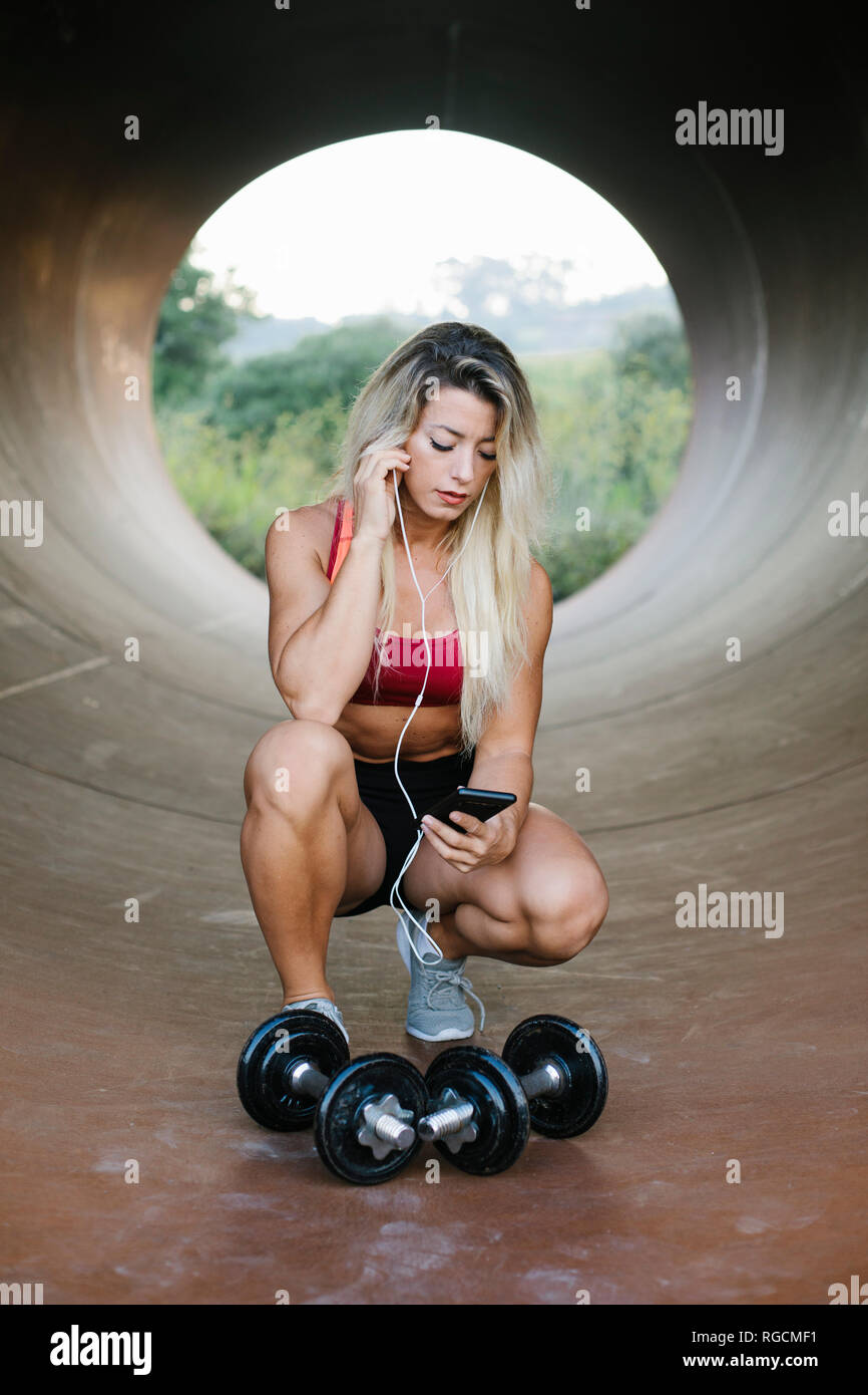 Athletic donna accovacciata all'interno di un tubo con manubri e auricolari guardando al telefono cellulare Foto Stock