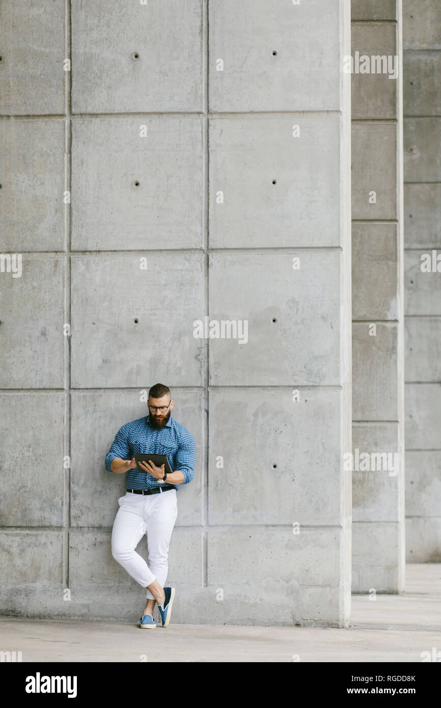 Tanga barbuto imprenditore indossando occhiali e plaid shirt appoggiata contro la parete utilizzando tablet Foto Stock