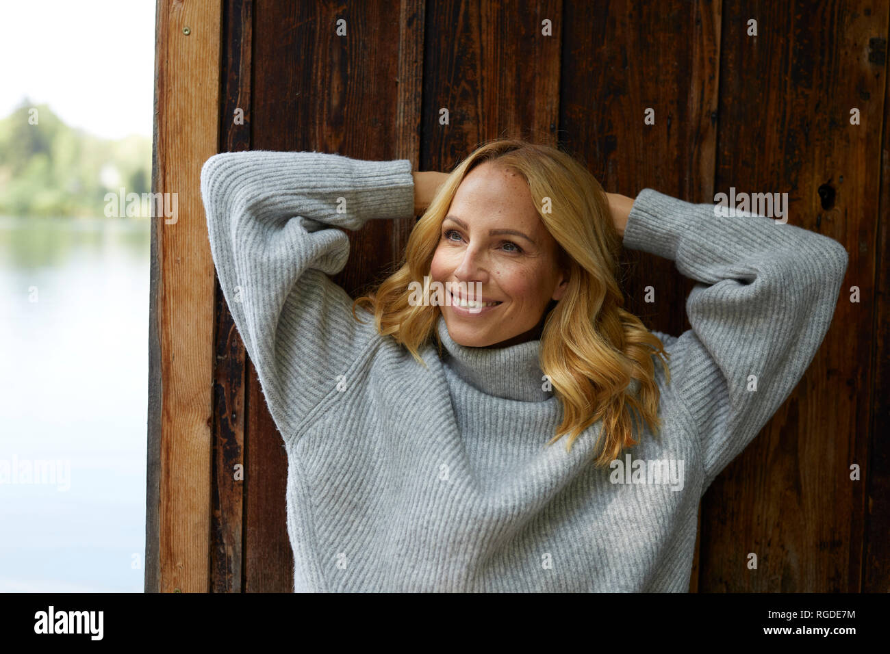 Sorridente donna bionda appoggiata contro la capanna in legno in un lago Foto Stock