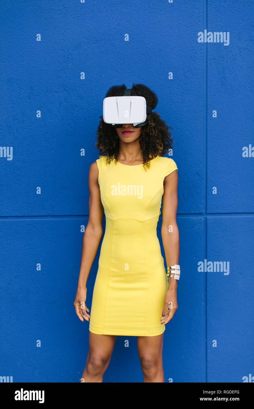 Donna in abito giallo indossando occhiali per realtà virtuale contro sfondo blu Foto Stock