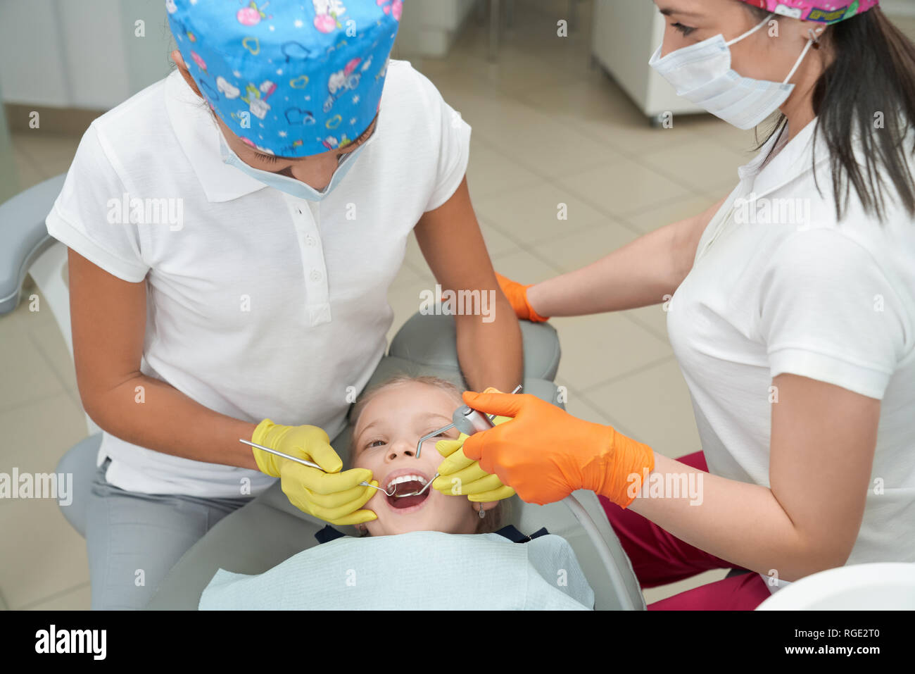 Due medici di controllo e trattamento di denti del piccolo paziente in  studio dentistico. Ragazza distesa sulla poltrona odontoiatrica con la  bocca aperta e guardando i dentisti positivo in maschere divertenti.  Concetto