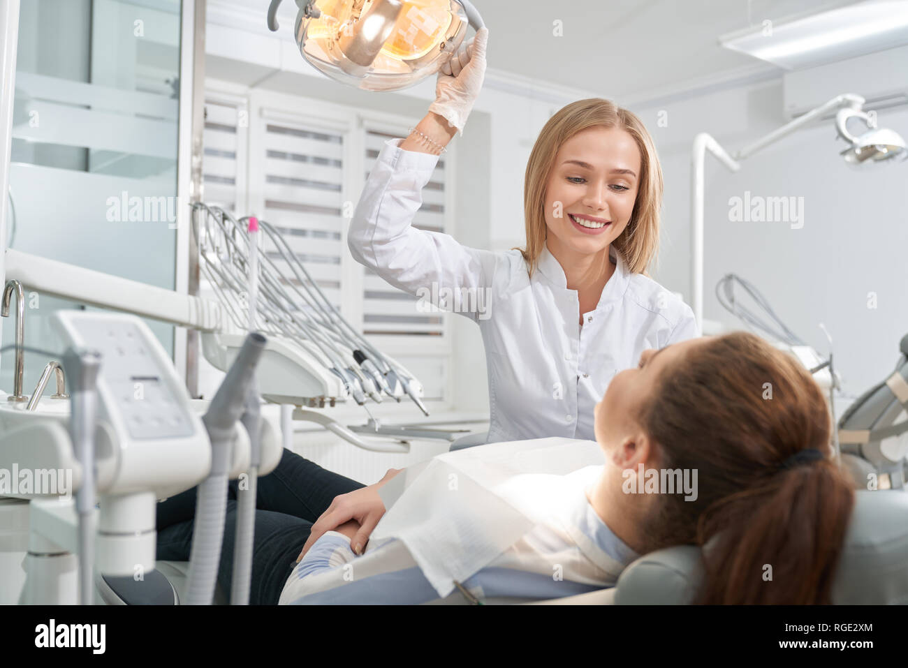 Paziente sdraiato sulla poltrona del dentista e la consultazione con il medico. Bella donna in bianco uniforme e guanti tenendo la mano su una speciale lampada, sorridente, guardando il client. Foto Stock