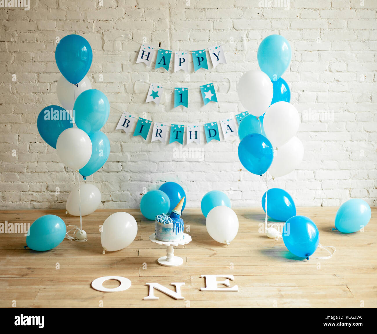 Decorazioni per un anno il compleanno con un sacco di blu e bianco palloncini, torta di festosa e scritte sul muro di mattoni e pavimento. Foto Stock