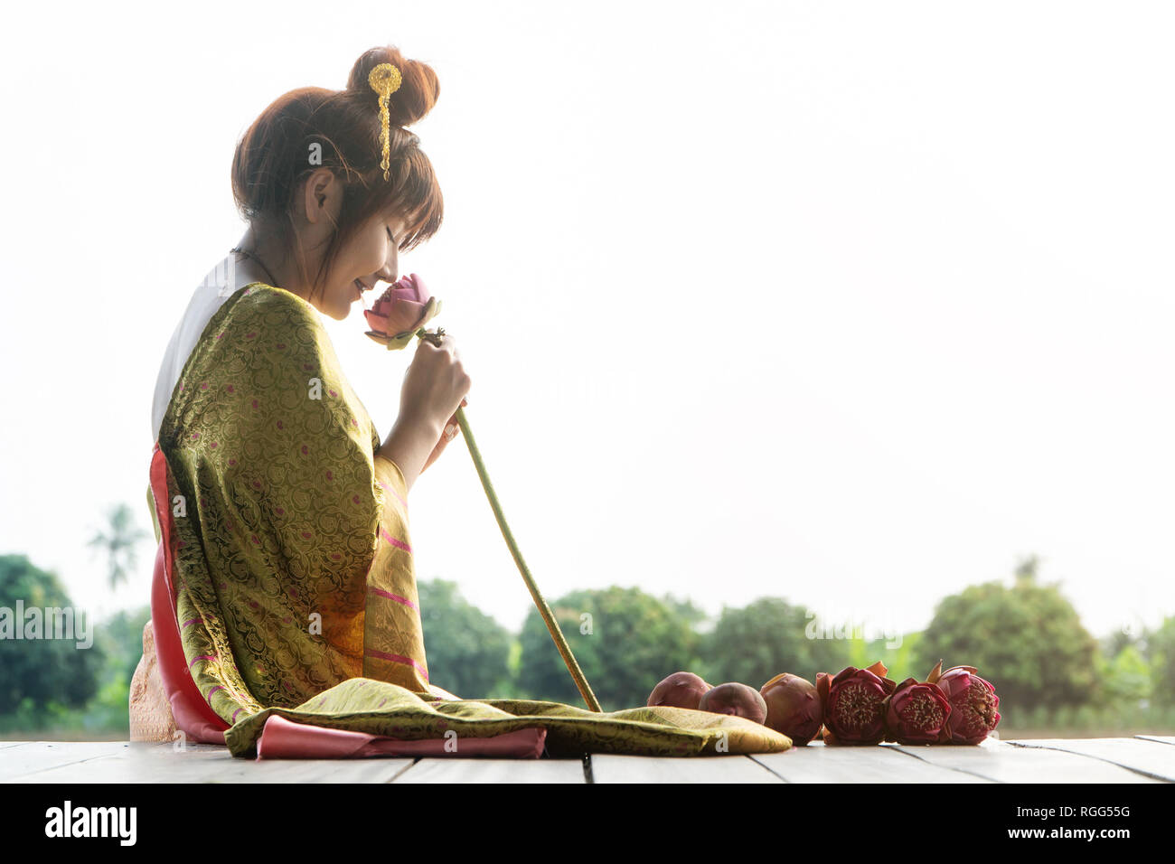 Bella asia donne indossando il tradizionale abito tailandese e seduto sul pavimento in legno. La sua mano è in possesso di fiore di loto e odore di fiori di colore rosa. Foto Stock