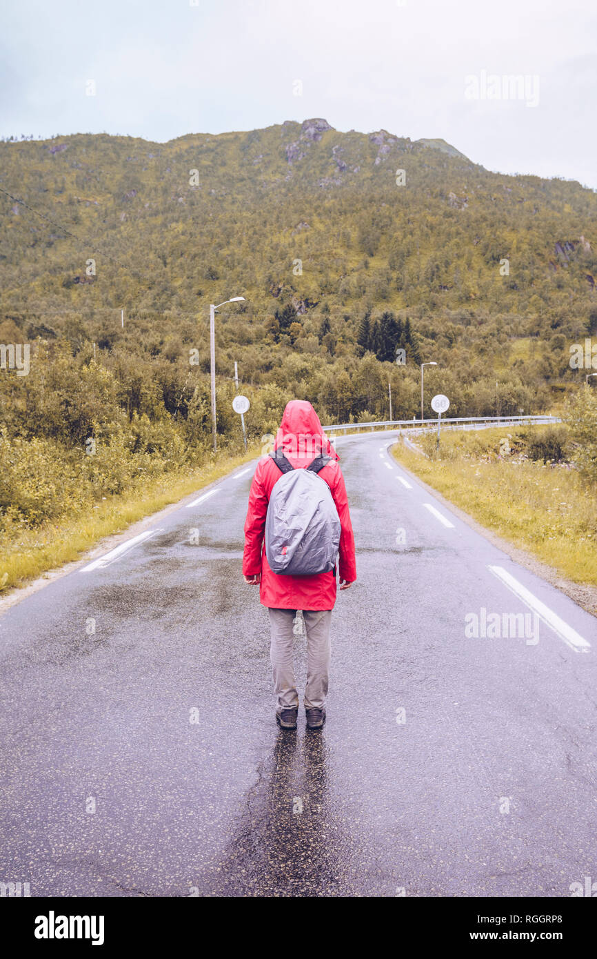 Norvegia Lofoten, vista posteriore dell uomo che indossa pioggia rossa acket e zainetto permanente sulla strada bagnata Foto Stock