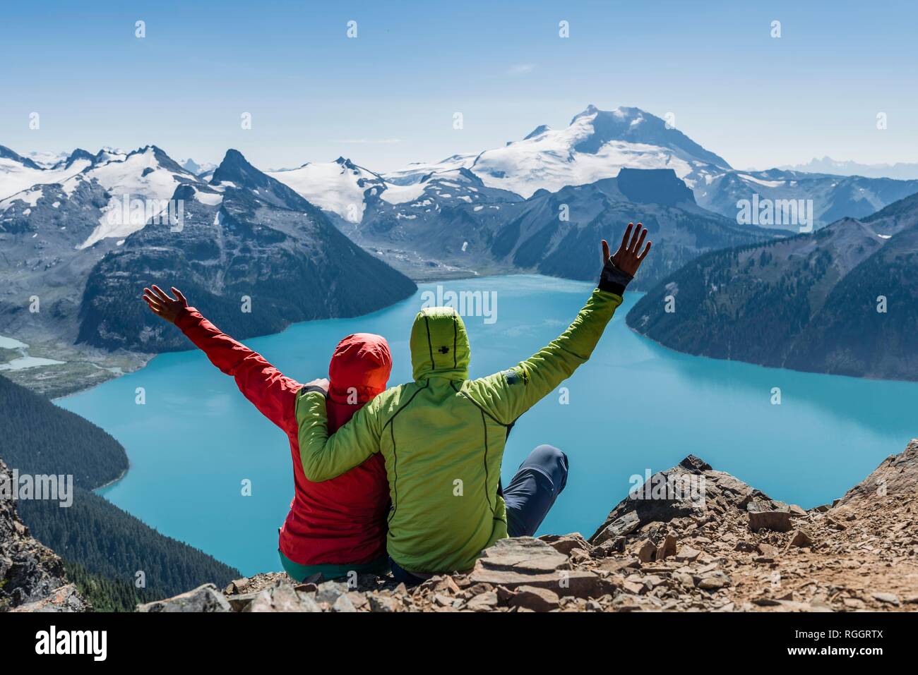 La vista dal crinale panoramico sentiero escursionistico, due escursionisti seduto su una roccia a bracci di estensione in aria, Lago di Garibaldi, turchese Foto Stock