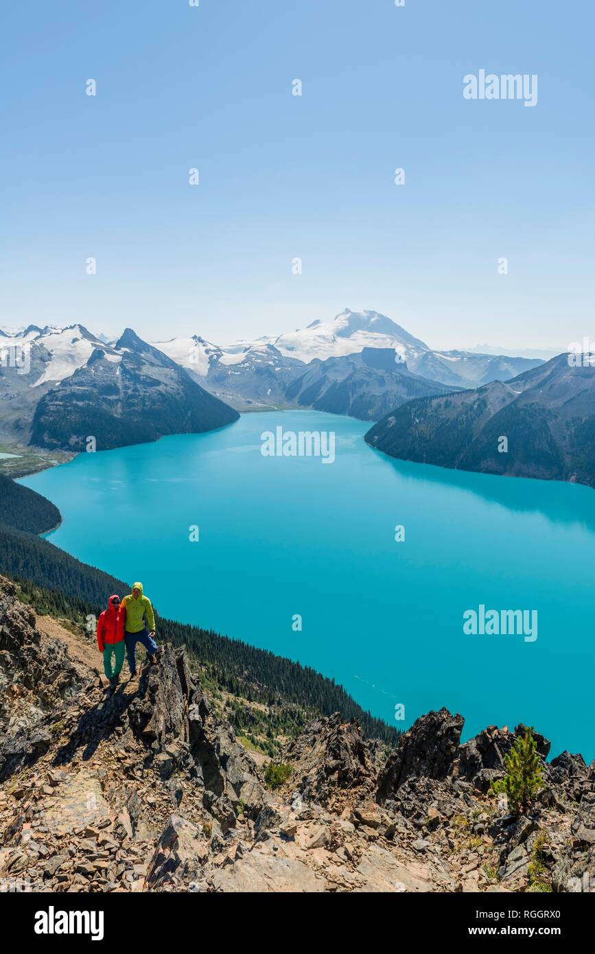Vista dal panorama Ridge trail, due escursionisti su una roccia, Lago di Garibaldi, turchese lago glaciale, Guardia di montagna e picco di inganno Foto Stock