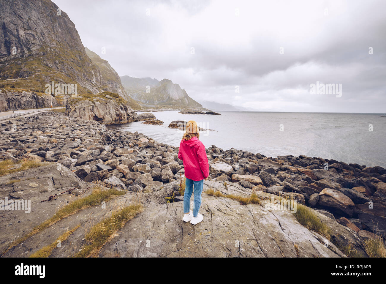 Norvegia Lofoten, vista posteriore della giovane donna indossa rainjacket in piedi su una roccia a guardare la distanza Foto Stock