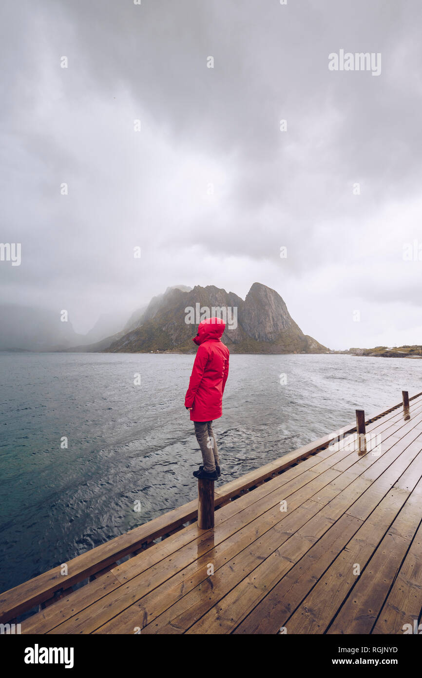 Norvegia Lofoten, Hamnoy, uomo rosso da indossare giacca pioggia in piedi sul paletto di legno guardando a distanza Foto Stock