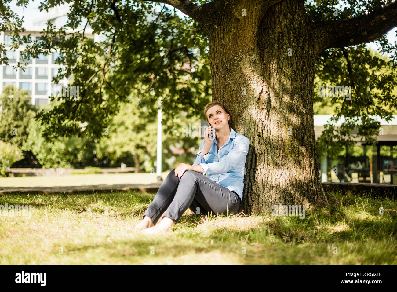 Donna seduta nel parco urbano appoggiata contro un albero parlando al cellulare Foto Stock