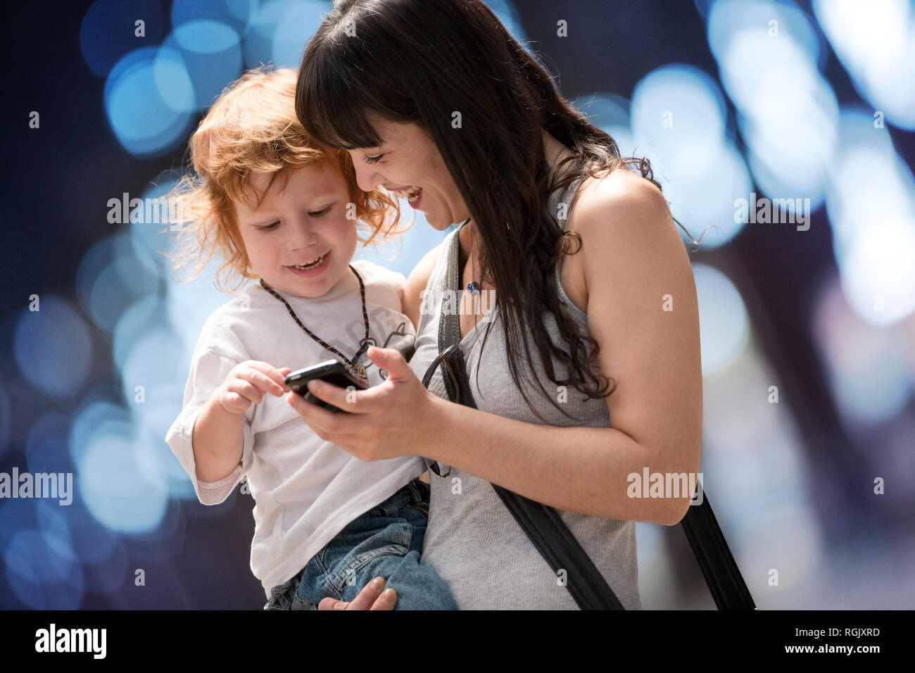 Madre porta il suo giovane figlio e hanno sorriso come ella gli mostra un telefono cellulare. Foto Stock