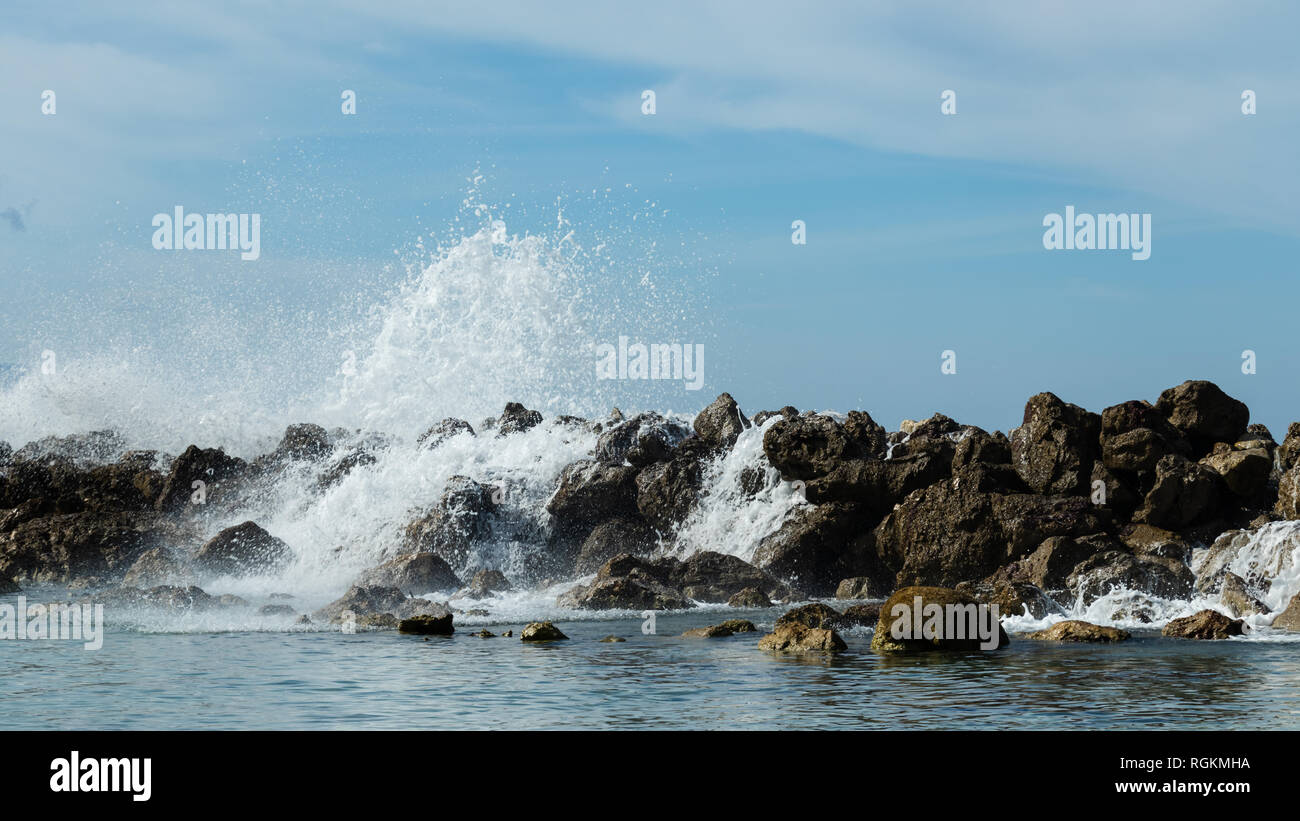 Onde infrangersi e versare sopra una parete di roccia formando una barriera protettiva per trattenere il potente mare. Calma acqua chiara con luminosi dei Caraibi estate cielo. Foto Stock