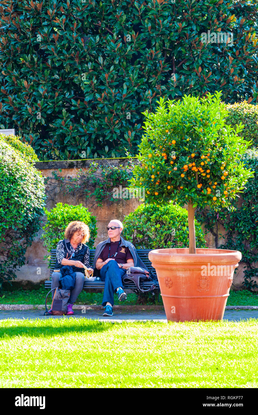 Vaticano, Roma, Italia - 16 Novembre 2018: due turisti, uomo e donna, giovane, seduto e appoggiato sul banco vicino all'albero di agrumi nel giardino quadrato Foto Stock