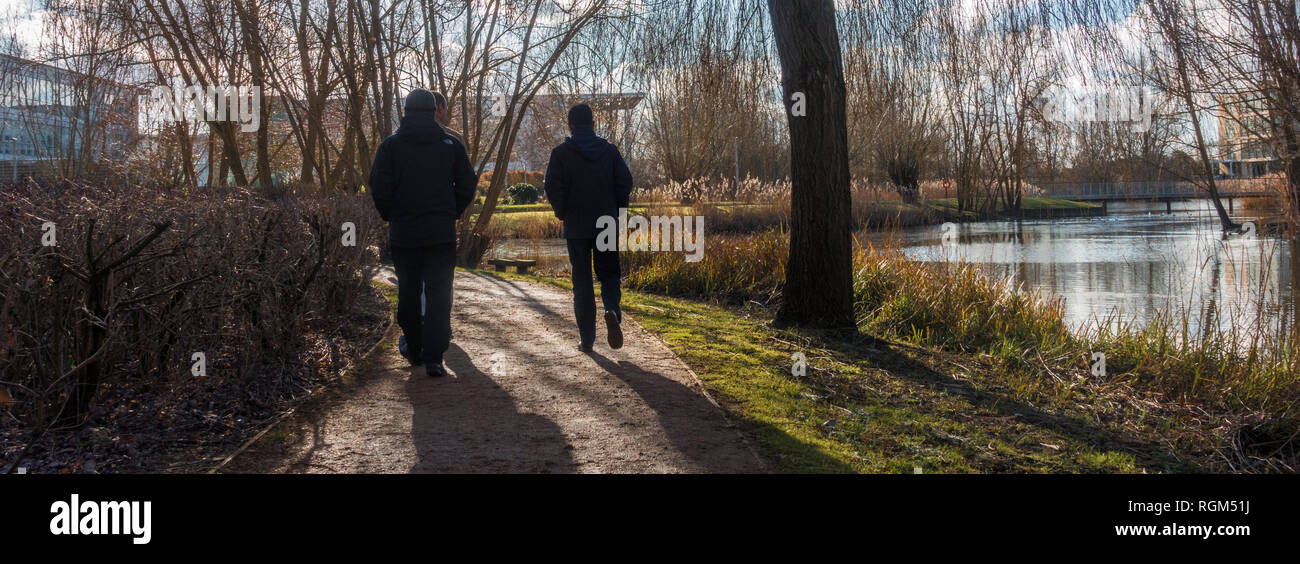 Un paio di uomini su una passeggiata intorno al lago nel mezzo di un parco commerciale a pranzo in inverno. Foto Stock