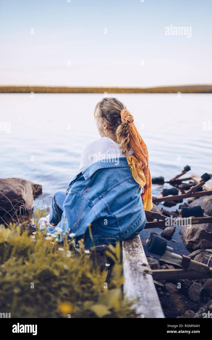 La Svezia, Lapponia, vista posteriore della giovane donna seduta a bordo d'acqua guardando a distanza Foto Stock
