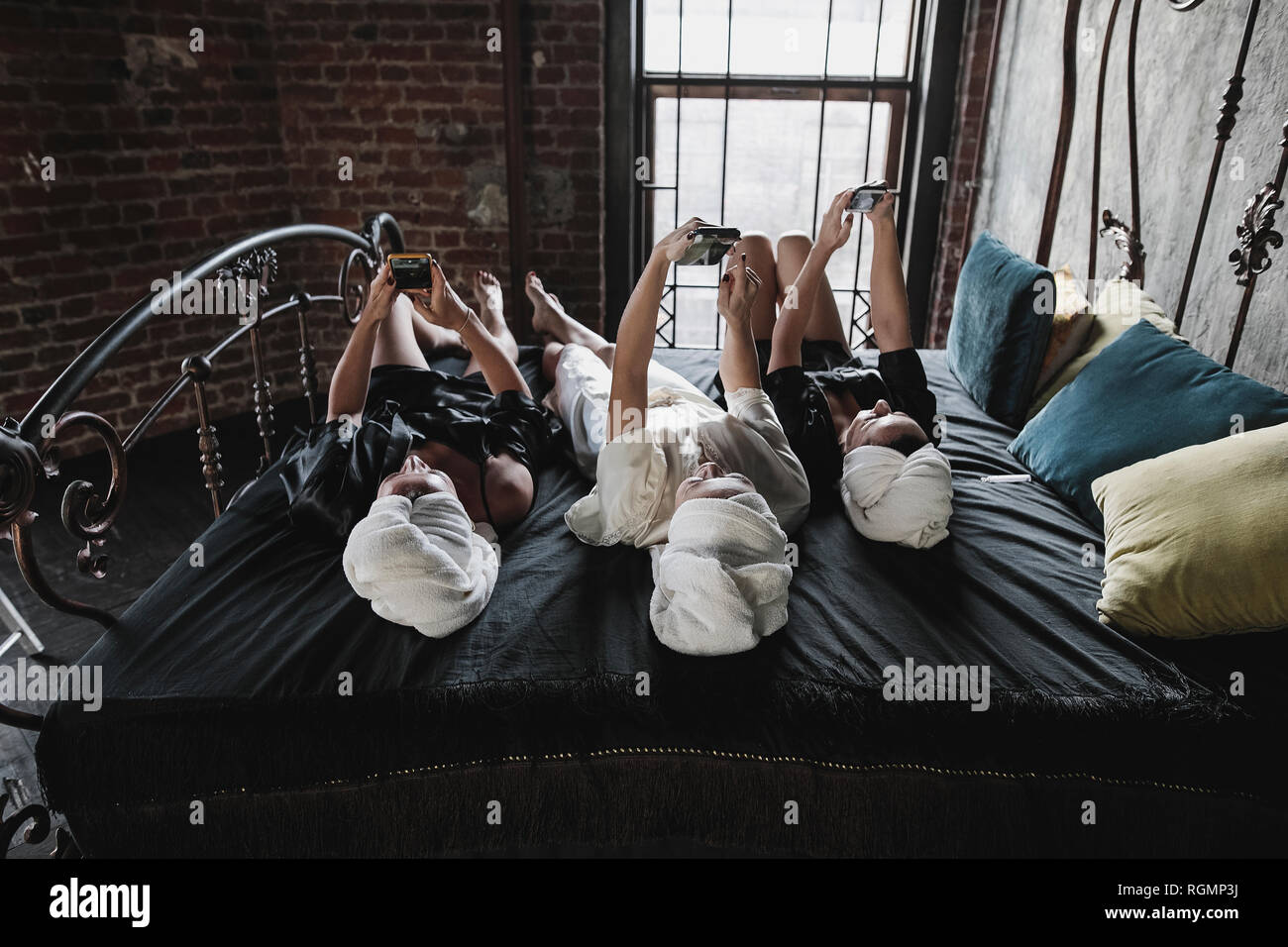 Tre donne con asciugamani intorno ai suoi capi giacente sul letto tenendo selfies Foto Stock