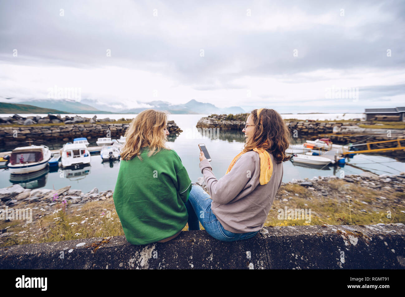 Norvegia, Senja, due giovani donna seduta in un piccolo porto con un telefono cellulare Foto Stock