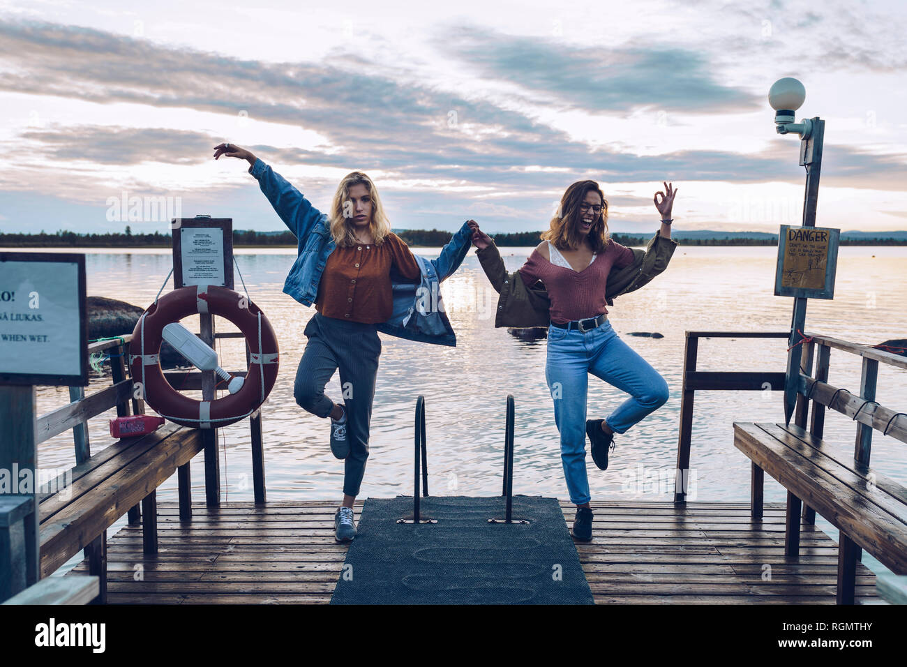 Due amiche in piedi su una gamba sola su un molo sul lago Inari, Finlandia Foto Stock