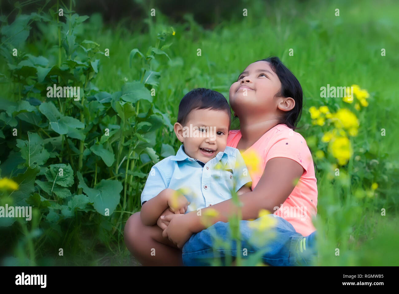 Una ragazza carina guardando il cielo, esprimendo la speranza mentre si tiene il suo bambino fratello nelle sue braccia in un campo esterno con fiori. Foto Stock