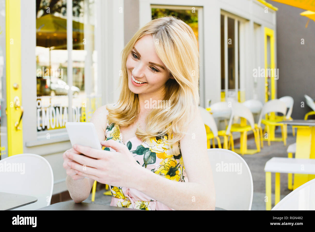 Una donna bionda guarda il suo cellulare e sorrisi a un cafe' all'aperto in estate Foto Stock