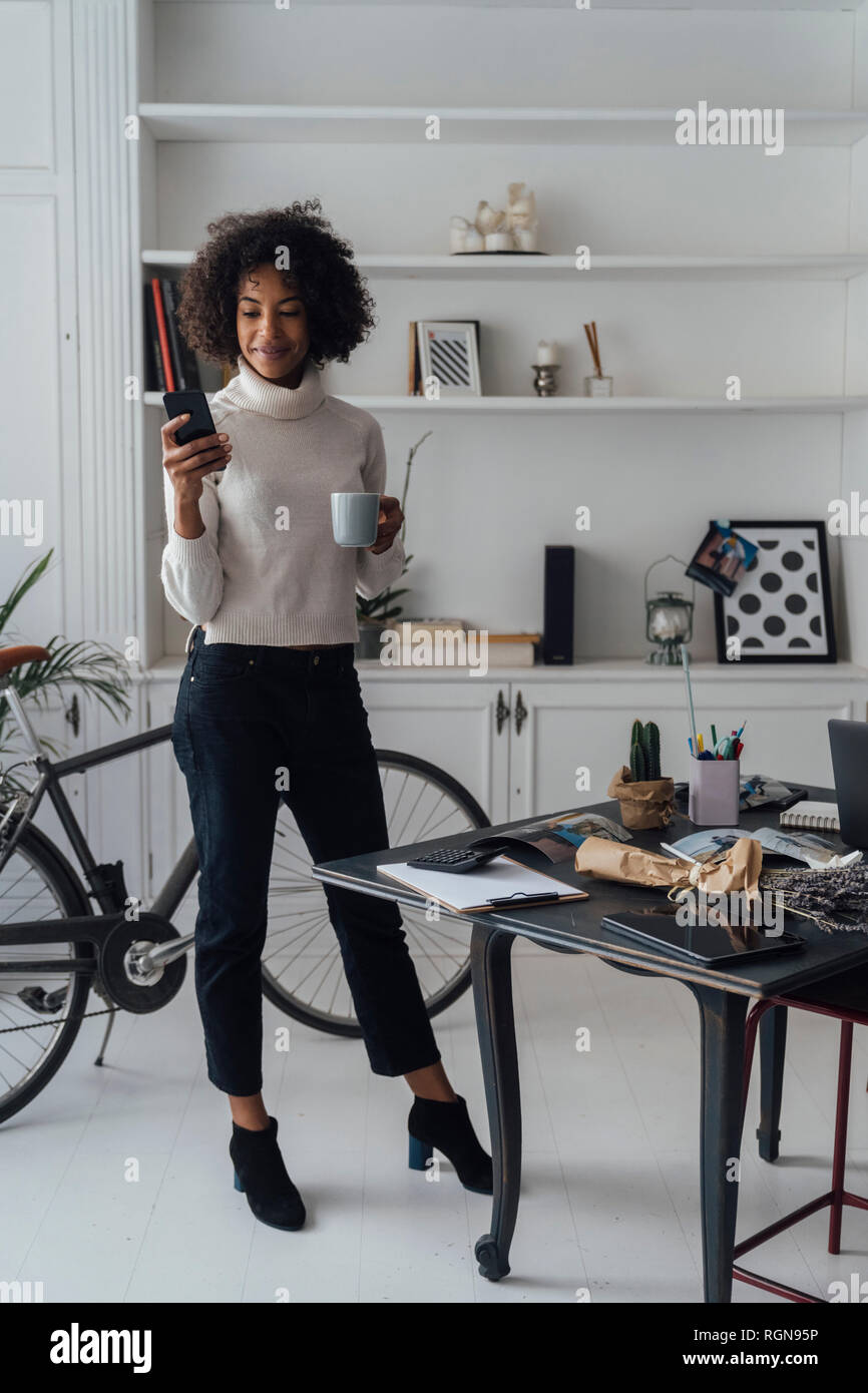 La metà degli adulti permanente del freelancer nel suo home office, utilizza lo smartphone, tenendo tazza da caffè Foto Stock