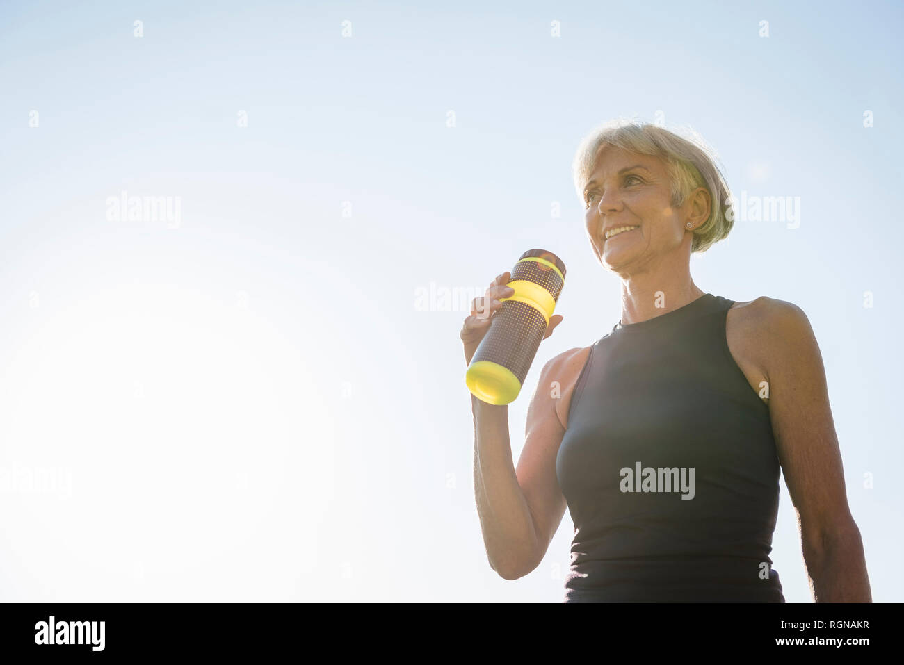 Sorridente sportive donna senior tenendo la bottiglia sotto il cielo blu Foto Stock