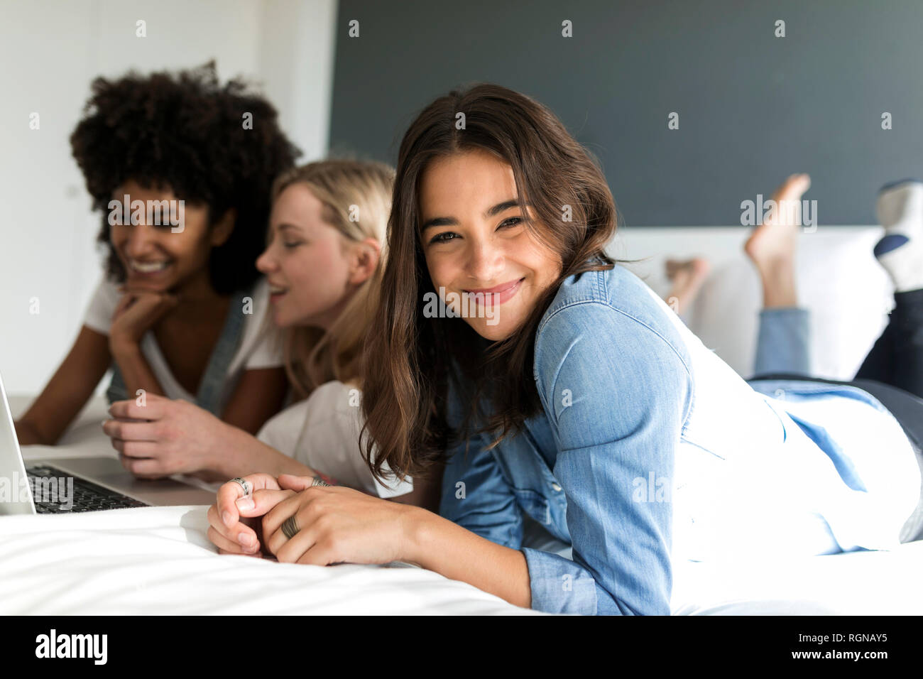 Ritratto di giovane sorridente donna sdraiata con amiche sul letto tramite computer portatile Foto Stock