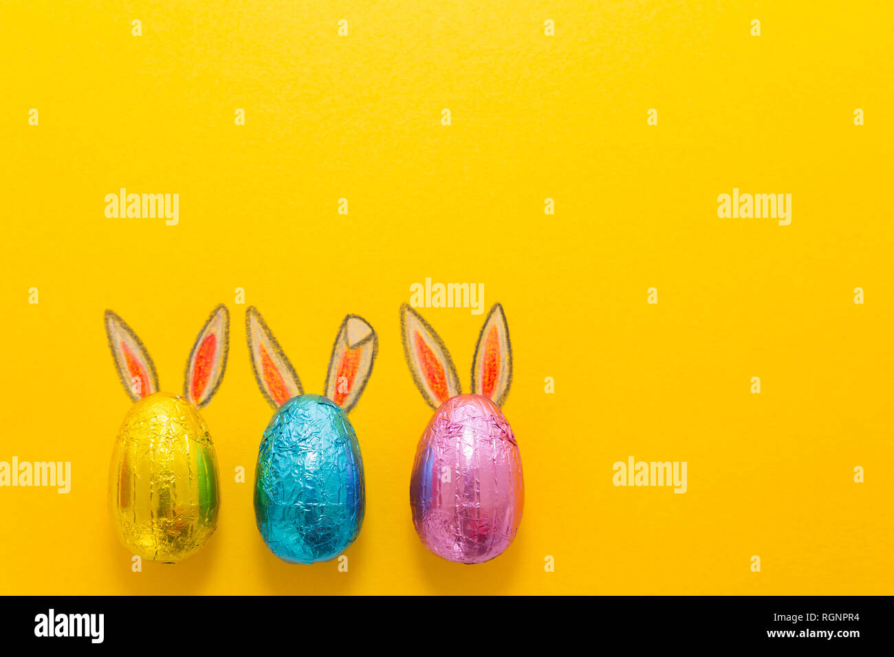 Tre uova di cioccolato colorato in lamina di alluminio verniciato con il coniglietto di pasqua le orecchie su sfondo giallo Foto Stock