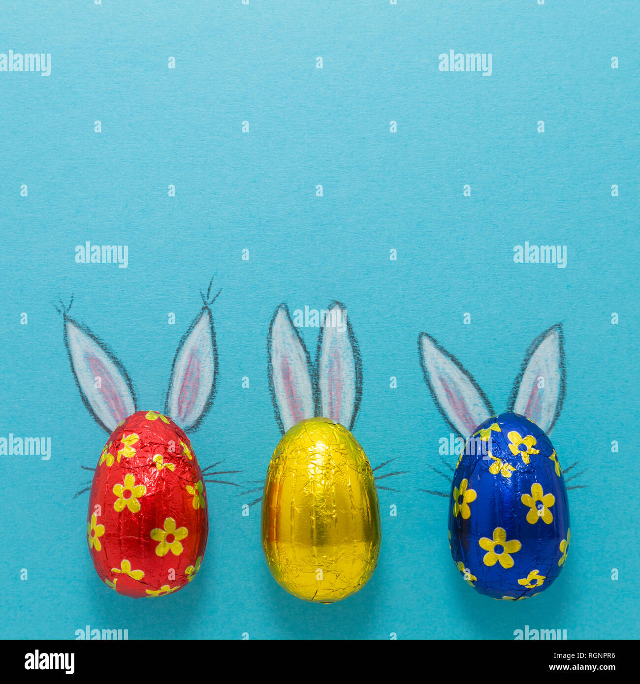 Tre uova di cioccolato colorato in lamina di alluminio verniciato con il coniglietto di pasqua le orecchie su sfondo blu Foto Stock