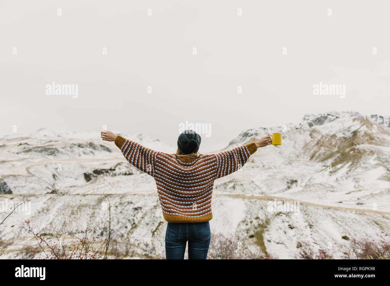 Vista posteriore del giovane lady in maglione e hat tenendo la tazza con le mani ai lati colline vicine nella neve in Pirenei Foto Stock