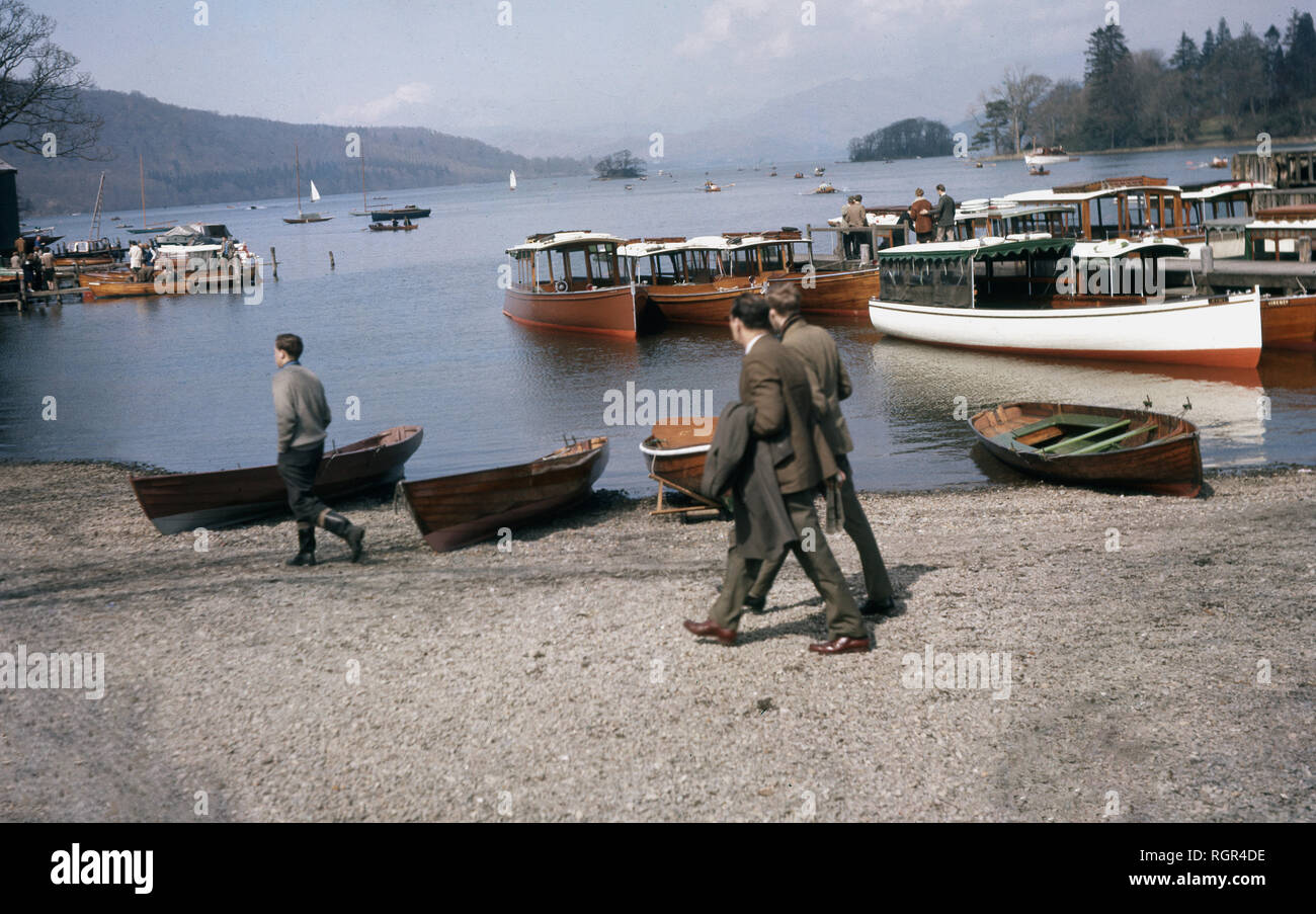 Degli anni Cinquanta, storico, gente camminare passato il turista in legno barche ormeggiate presso la riva di Rydal acqua, nel distretto del lago, Cumbria, Inghilterra, Regno Unito. Sebbene sia uno dei più piccoli laghi della zona, Rydal acqua è molto popolare ed è stato un favorito del poeta William Wordsworth, che hanno portato una casa all'estremità settentrionale del lago, "Muschio Bianco Casa". Foto Stock