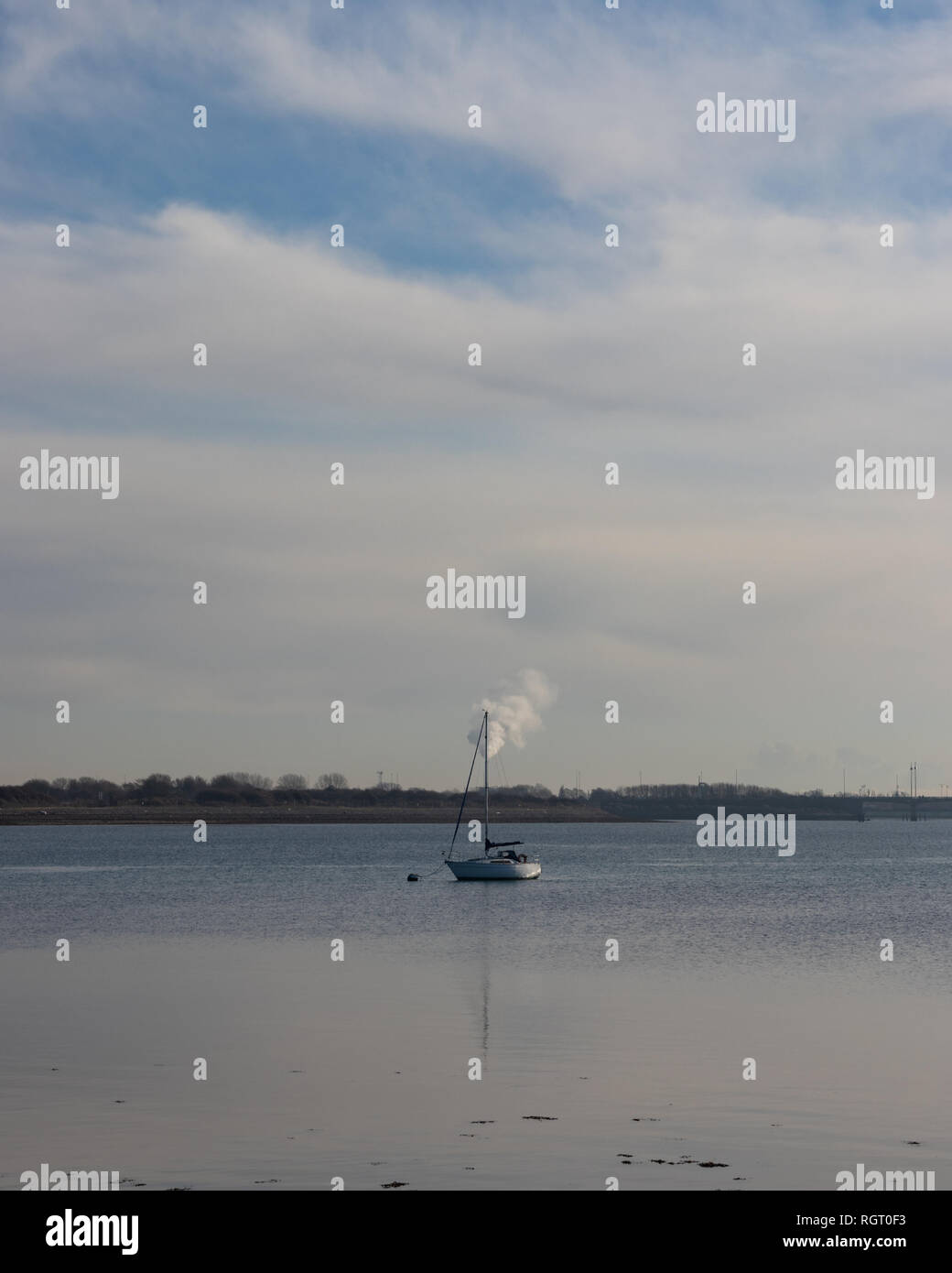 Barca a vela ancorata in acqua calma vicino alla riva con un inceneritore in background Foto Stock