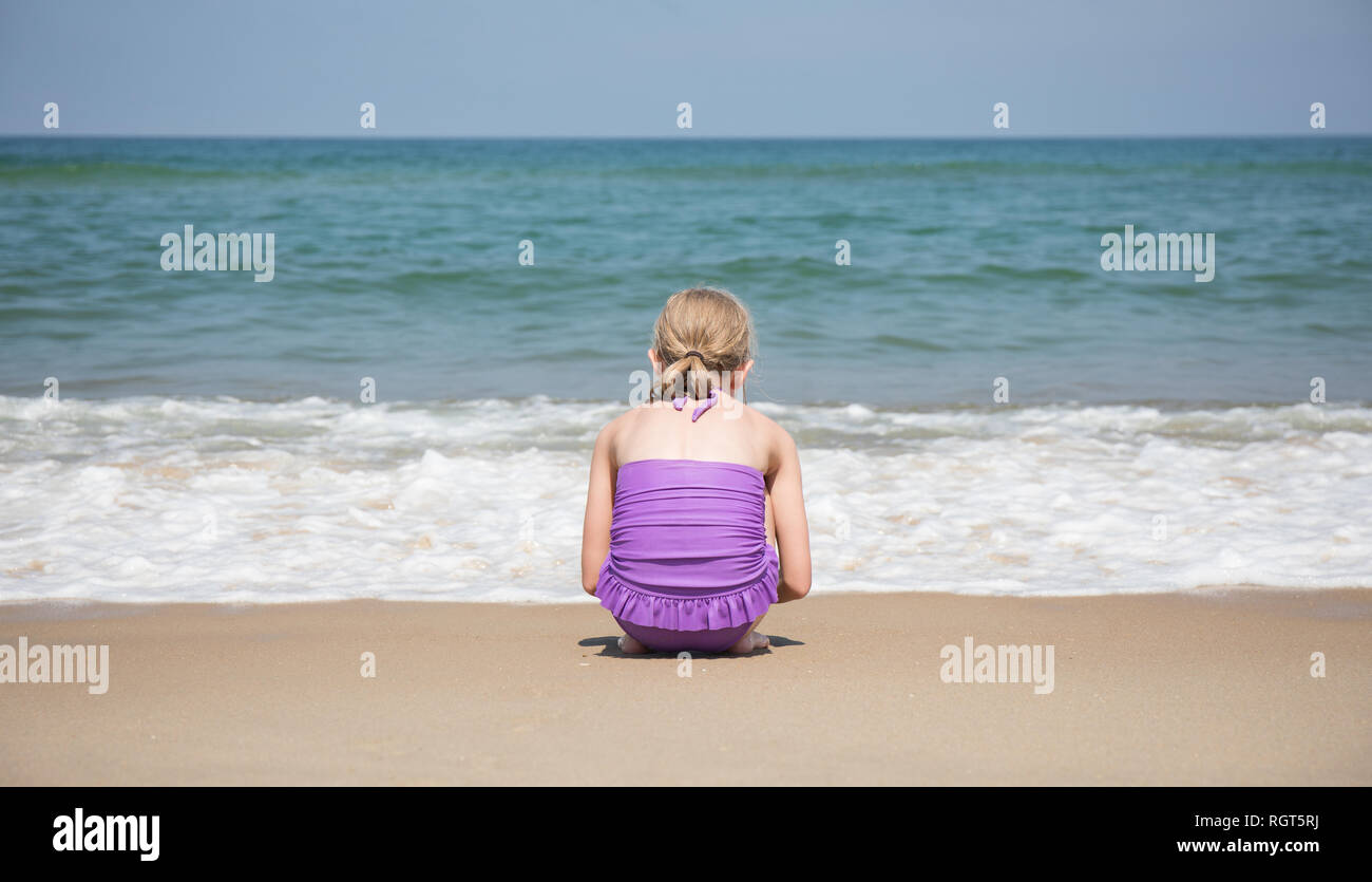 Un 9 anno vecchia ragazza accovacciato di fronte alle onde in arrivo a riva sulla spiaggia. Foto Stock