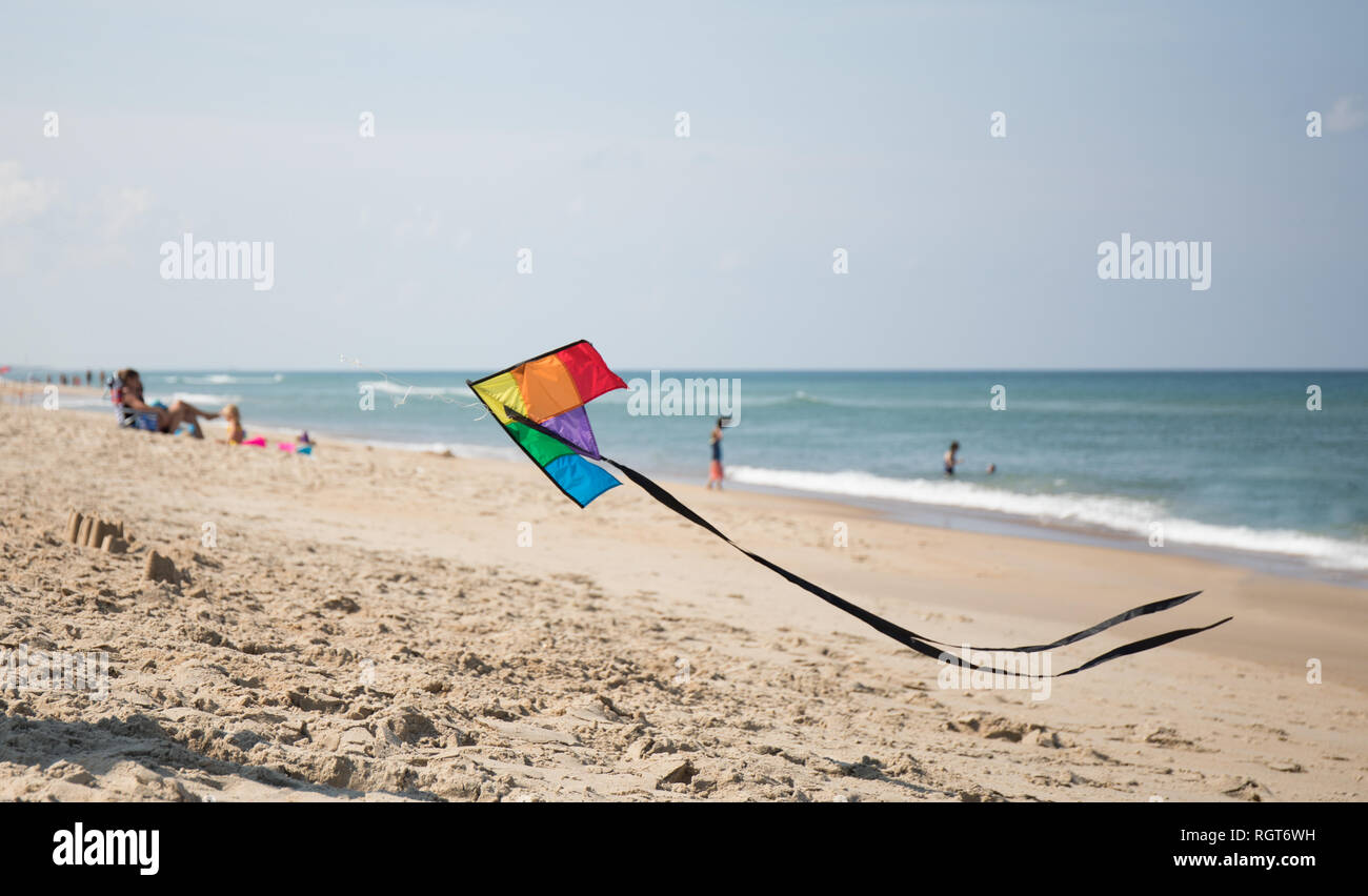 Un arcobaleno colorato aquilone volando a bassa quota sopra la spiaggia con le famiglie e vacanzieri in background. Foto Stock