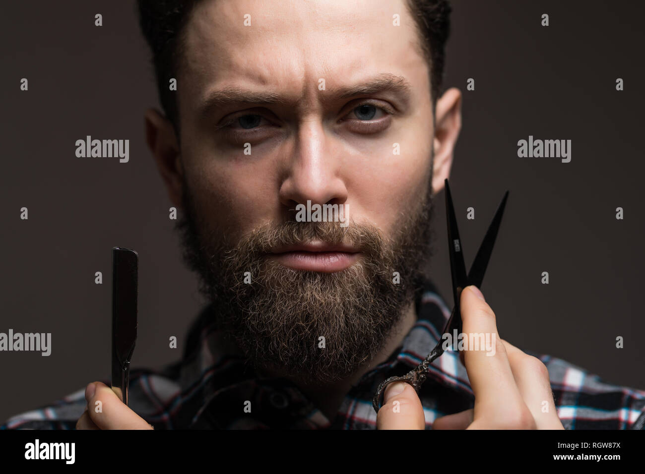 Ritratto di uomo barbuto con forbici da barbiere e il rasoio in piedi isolato su sfondo grigio Foto Stock