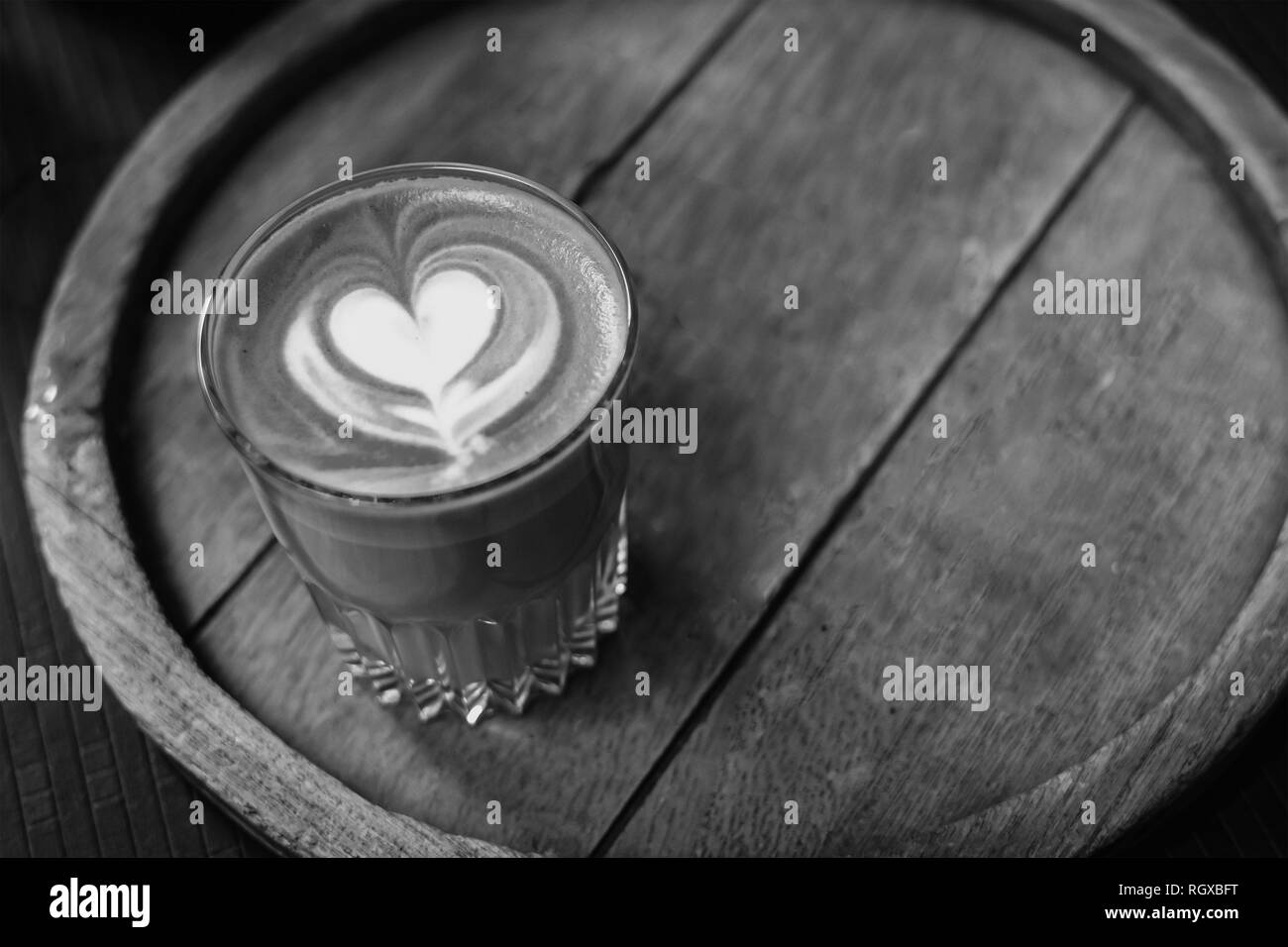 Una tazza di caffè con un cuore schiuma di latte art sulla parte superiore, seduto su un vassoio in legno, in bianco e nero, monocromatiche. Foto Stock
