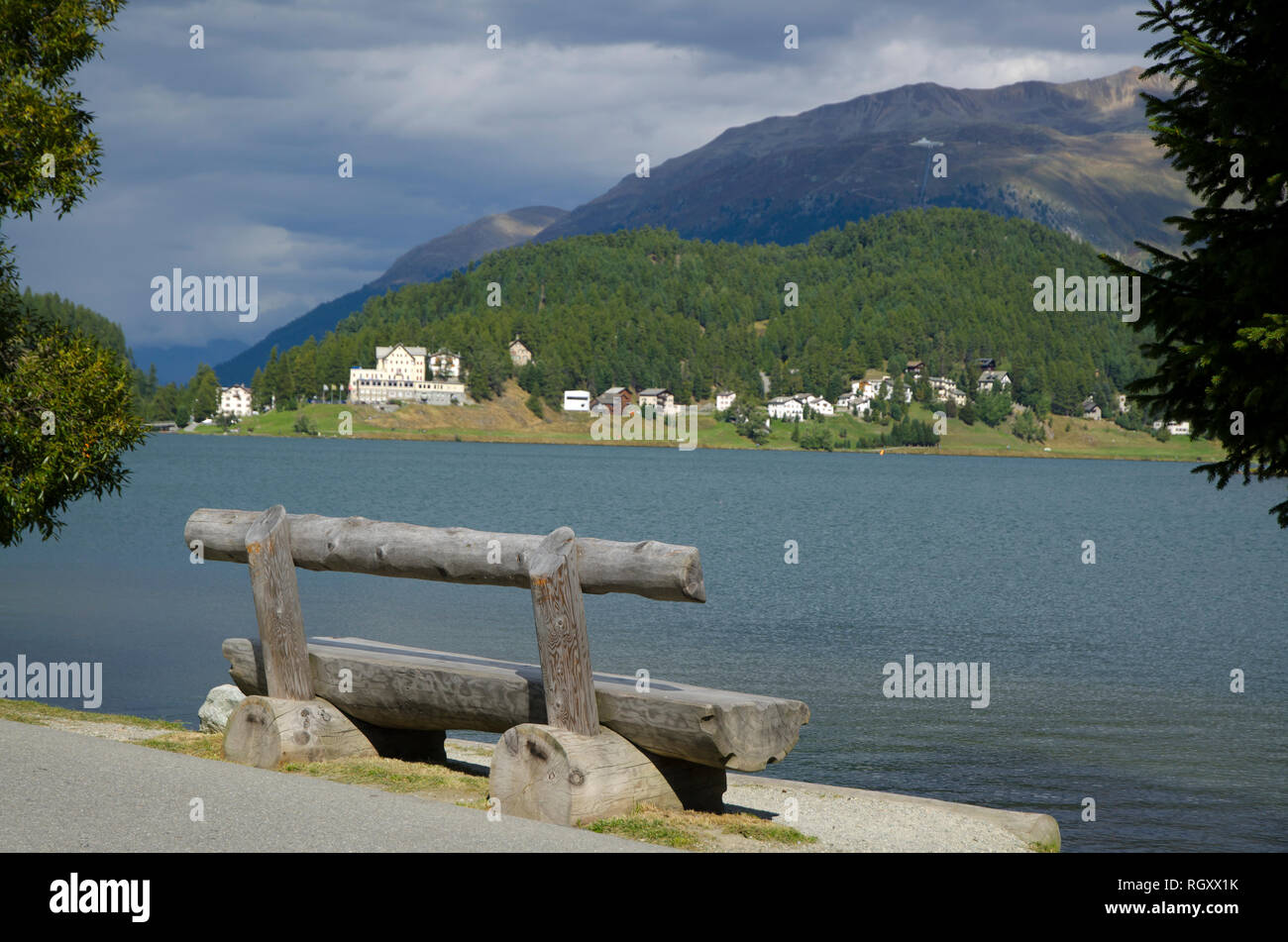 Panca di legno e gli alberi di St Moritz il lago con la montagna e le nuvole grigio in una giornata di sole in Svizzera. Foto Stock