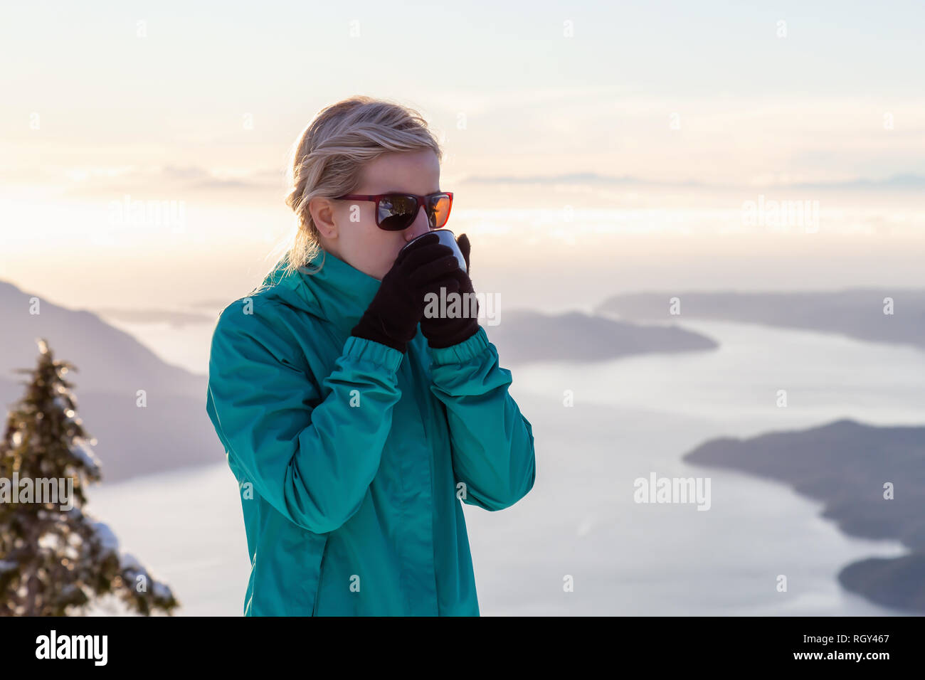 Giovane ragazza caucasica bere bevande calde sulla cima di una montagna durante un bellissimo tramonto in inverno. Prese su Mnt Harvey, vicino a Vancouver, BC, Canada. Foto Stock