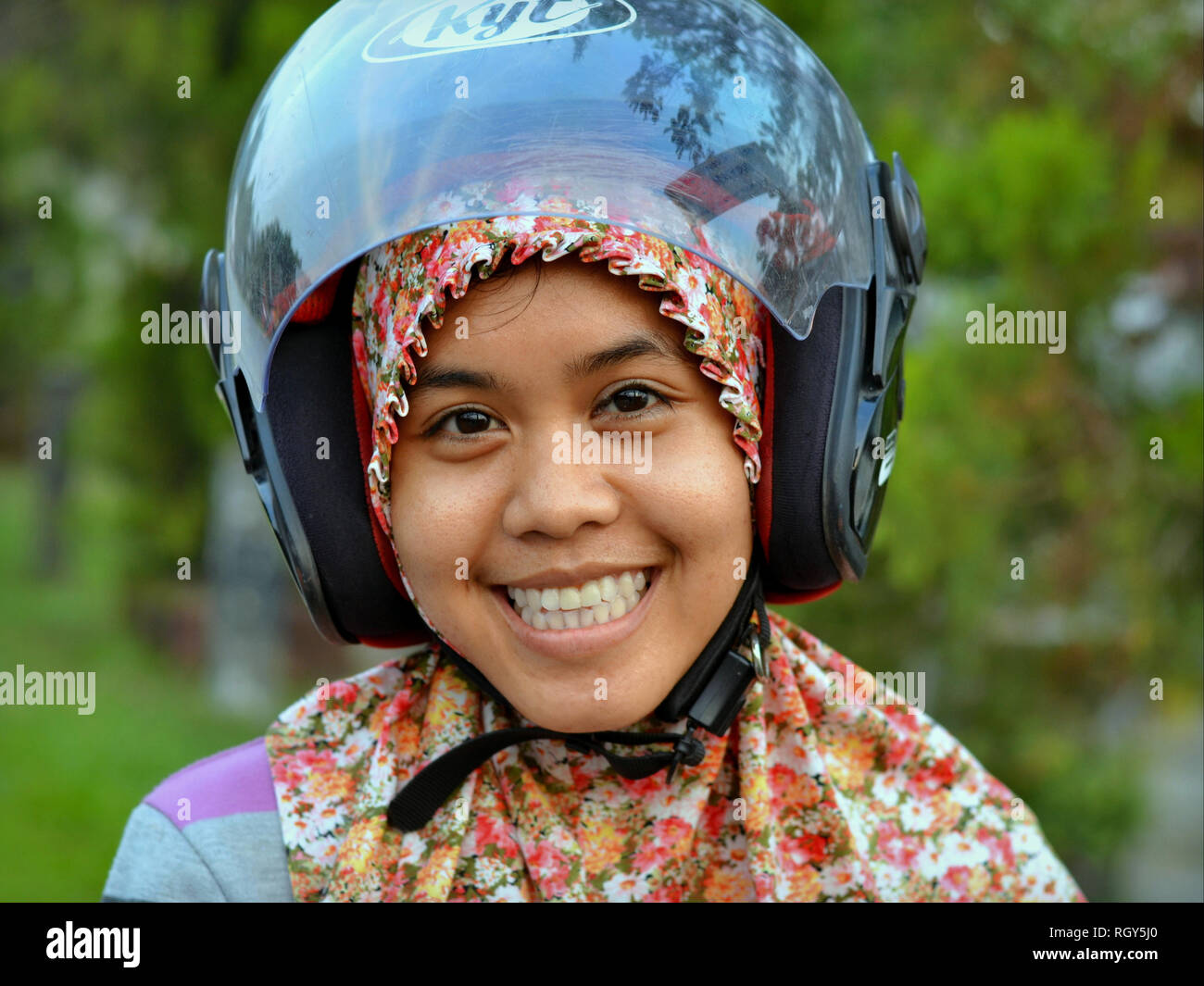 Giovane indonesiano donna dal Kalimantan indossa un moderno motociclo casco oltre il tradizionale suo hijab e sorrisi per la fotocamera. Foto Stock