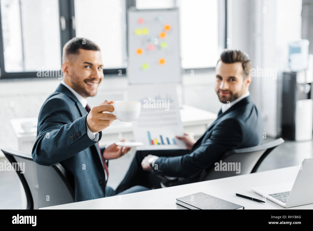 Messa a fuoco selettiva del bicchiere con la bevanda in mano di allegro imprenditore vicino collaboratore tenendo la carta con grafici Foto Stock