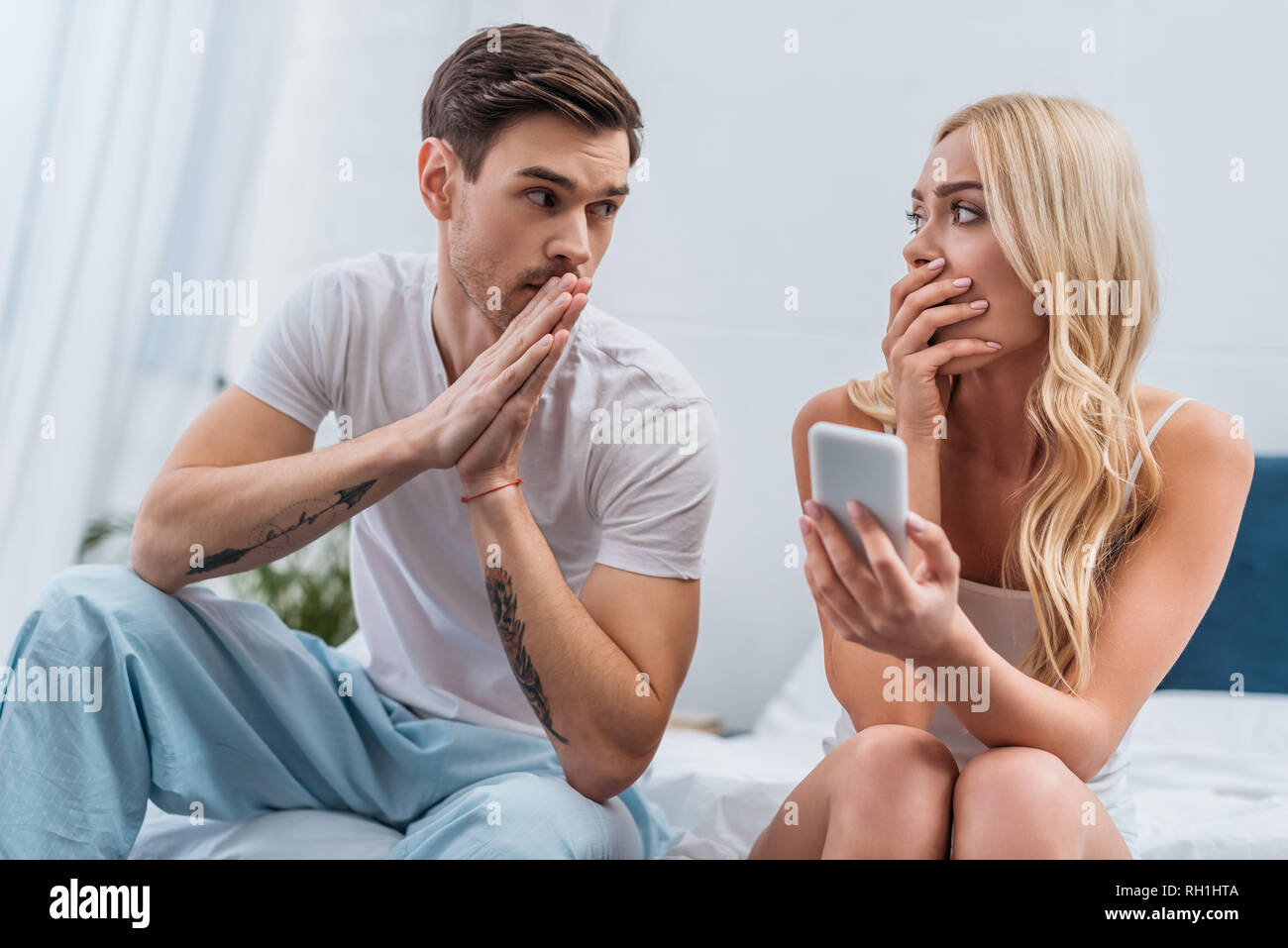 Scioccato donna tenendo lo smartphone e guardando confuso marito seduta sul letto e la diffidenza, di concetto Foto Stock