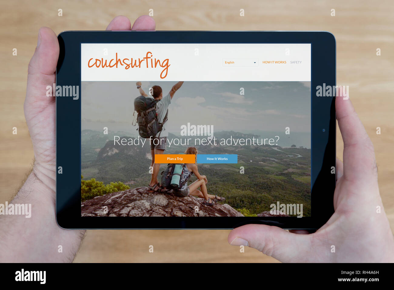 Un uomo guarda al Couchsurfing sito sul suo iPad dispositivo tablet, sparato contro un tavolo di legno top sfondo (solo uso editoriale) Foto Stock