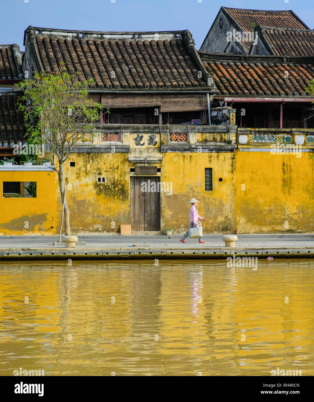 Locale donna vietnamita con riso hat a piedi nella parte anteriore del case tradizionali con elementi ornamentali pareti gialle e tetti di tegole Foto Stock