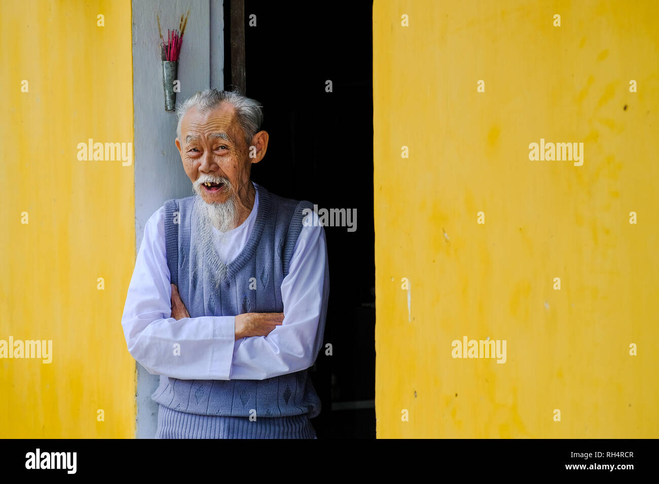 Saggio vietnamita vecchio uomo sorridente e gente accogliente a casa sua con pareti gialle in Hoi An, Vietnam. Foto Stock