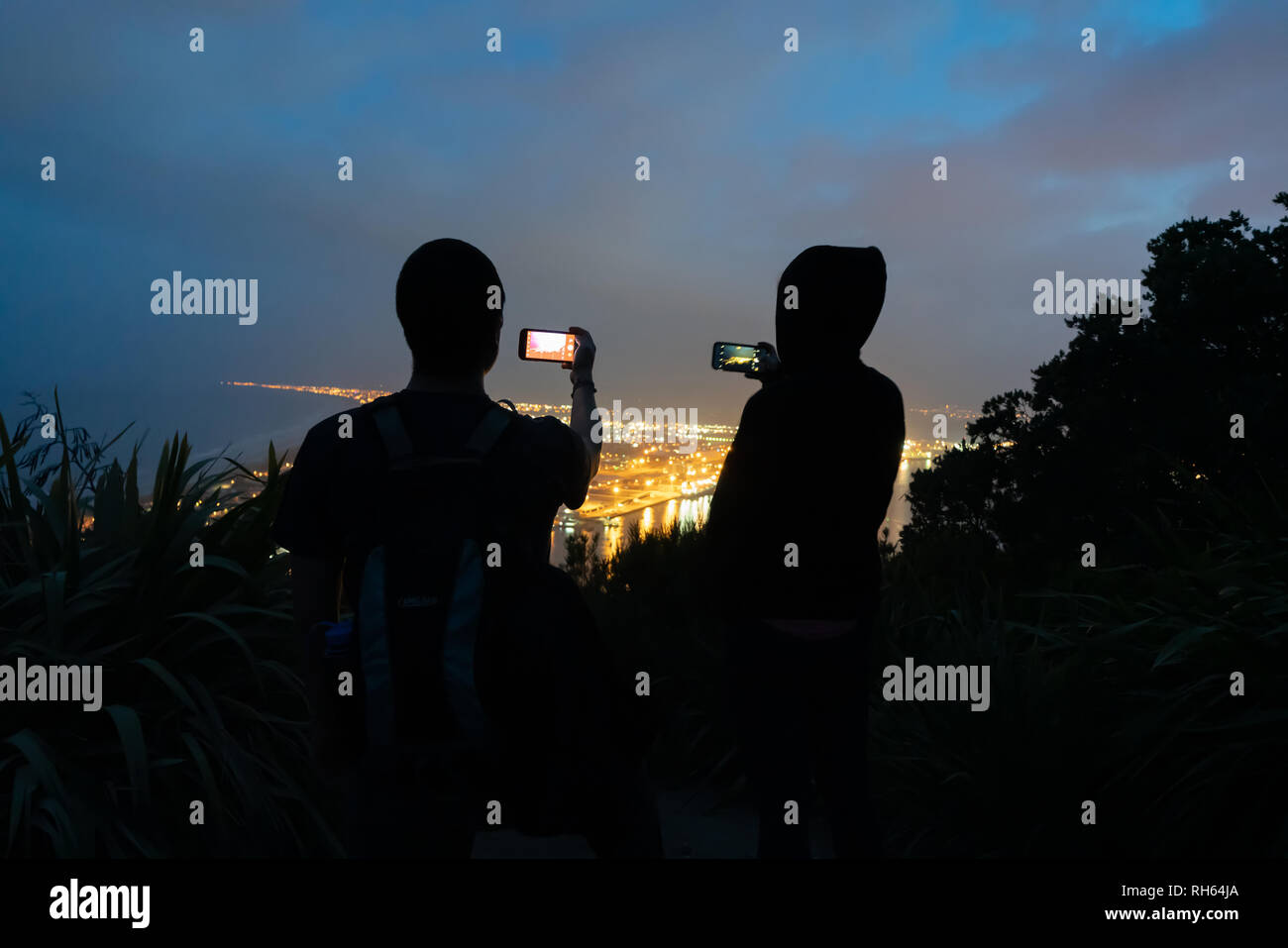 Immagine granulosa due irriconoscibile turisti in silhouette utilizzando telecamere su dispositivi mobili e telefoni per fotografia urban di notte le luci dalla parte superiore del punto di riferimento Mo Foto Stock