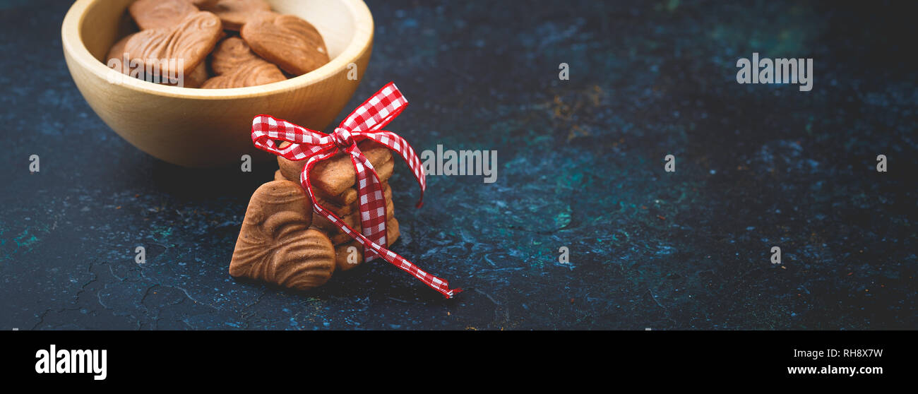 Pila di zenzero a forma di cuore i cookie per il giorno di San Valentino con il bianco-nastro rosso su sfondo blu scuro. Spazio per il testo. Lungo banner largo Foto Stock