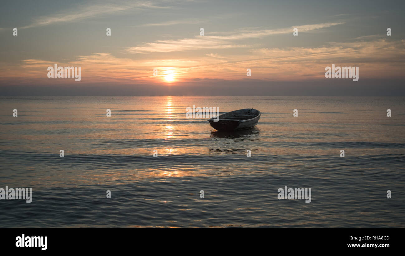 Una barca vuota circondata da piccole ondulazioni del Mar Baltico nella luce calda di lontani Rising Sun su una tranquilla mattina ancora. Foto Stock