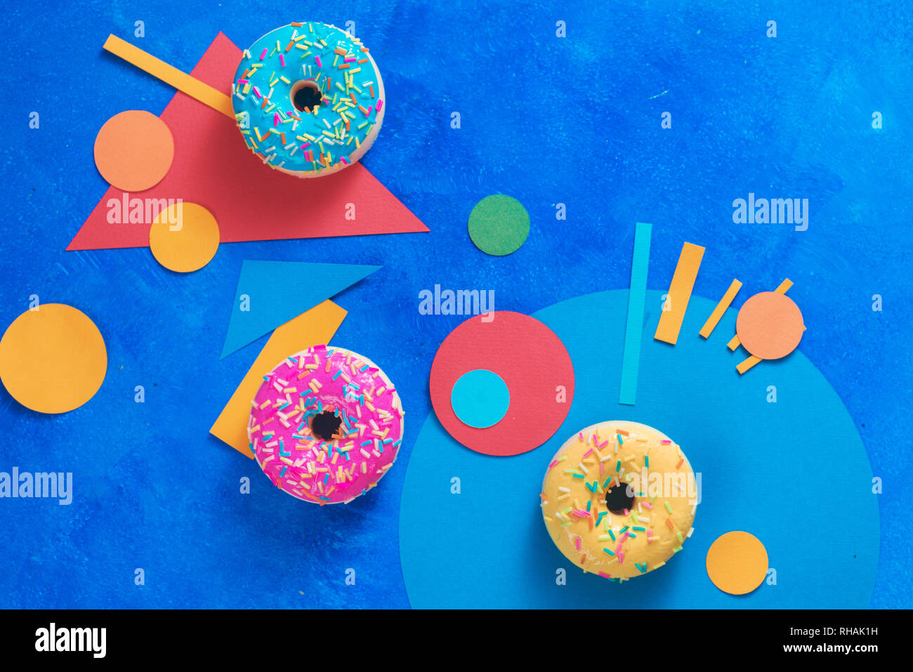 Le ciambelle di diversi colori su uno sfondo blu con forme geometriche. Il blocco di colore piatto alimentare lay con copia spazio. Foto Stock