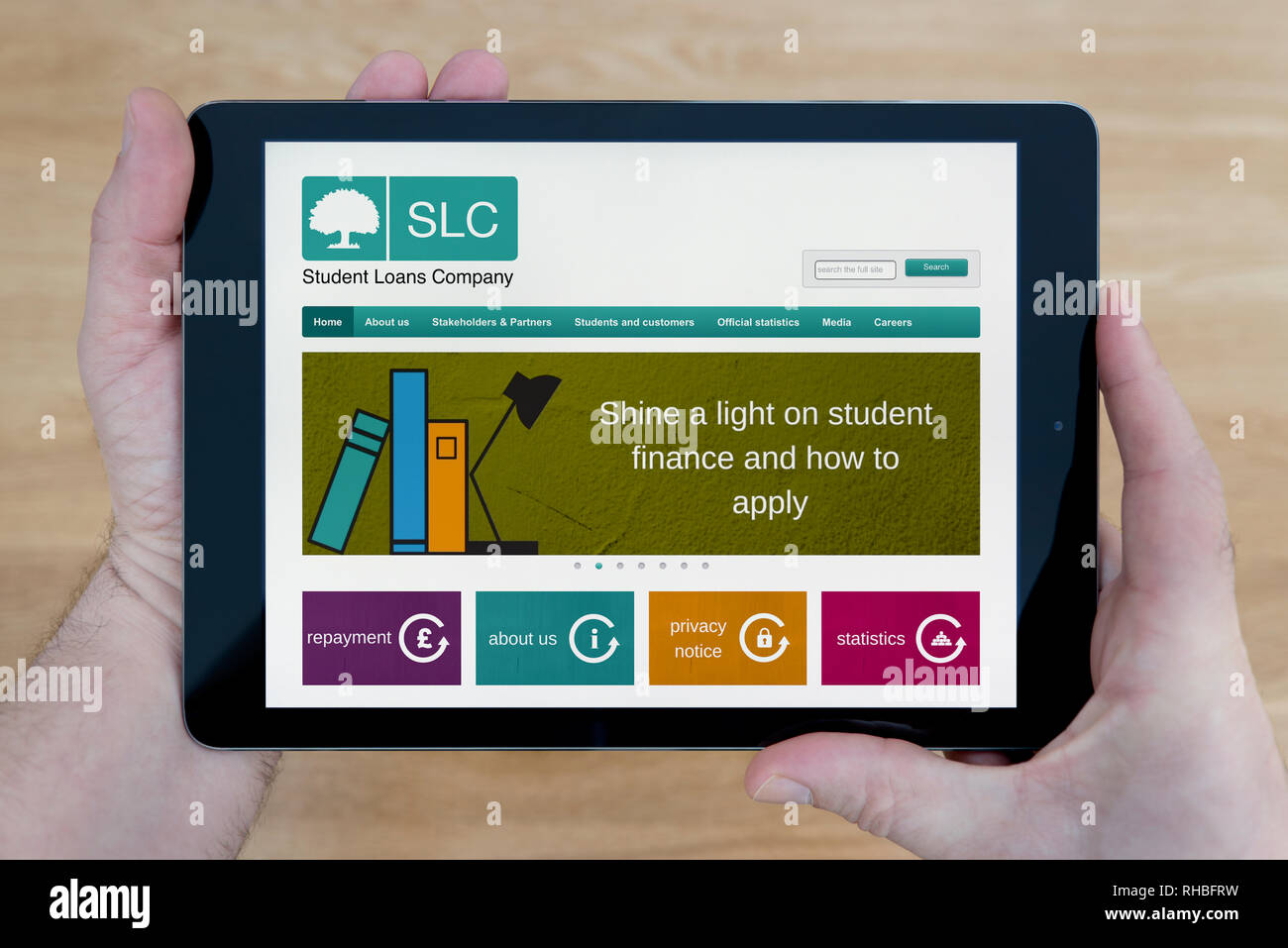 Un uomo guarda al Student Loans Company (SLC) Sito web sul suo iPad dispositivo tablet, sparato contro un tavolo di legno top sfondo (solo uso editoriale). Foto Stock